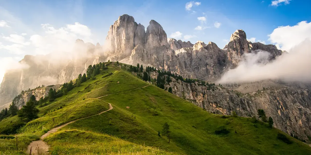 Sentiero serpeggiante su un prato verdeggiante ai piedi di un imponente gruppo montuoso tra Veneto e Trentino Alto Adige, con nuvole che accarezzano le cime rocciose.