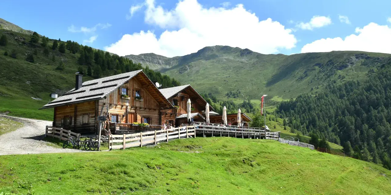Rifugio in legno con tavoli all'aperto, ideale per un giro delle malghe in Val di Non. Montagne verdi e cielo azzurro.
