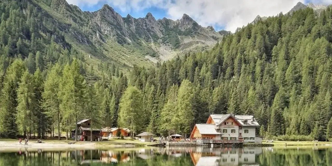 Riflessi sul lago alpino nella piana di Nambino, con boschi di conifere e montagne sullo sfondo.