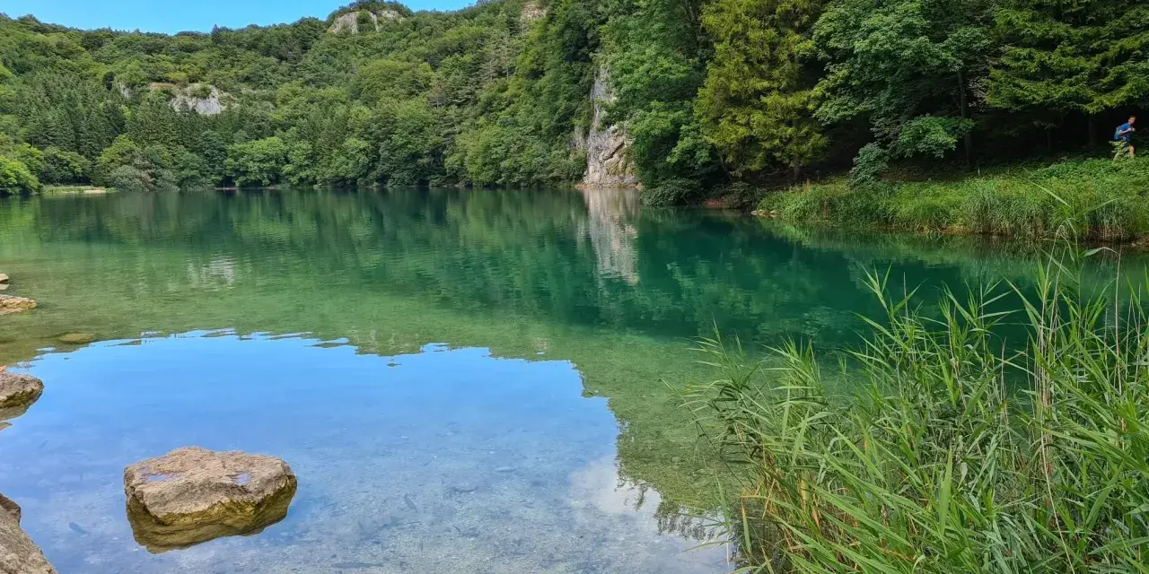 Acque cristalline del lago di Lavis riflettono la rigogliosa vegetazione. Rocce affiorano, un corridore si allena lungo la riva.