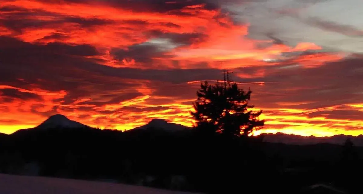 Un infuocato tramonto sulle Dolomiti, con nuvole rosse e arancioni che illuminano il cielo sopra le sagome scure delle montagne e di un pino solitario.