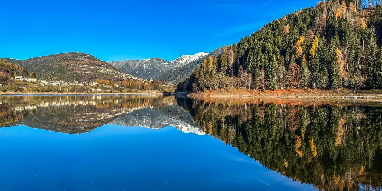 Il lago delle Piazze riflette montagne innevate, boschi autunnali e un villaggio sotto un cielo azzurro.
