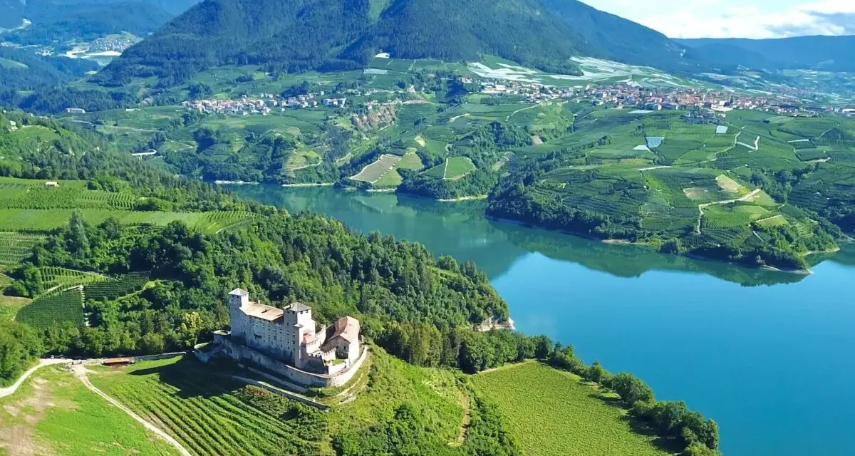 Castello medievale su una collina verdeggiante, con vigneti a terrazze e un lago azzurro nel **Val di Non**.