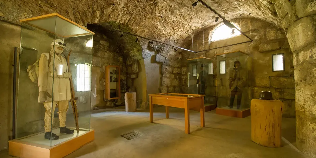Interno di un museo in pietra con manichini in uniformi storiche, uno dei quali indossa un abito chiaro e porta un fucile, evocando un forte strino di battaglia.