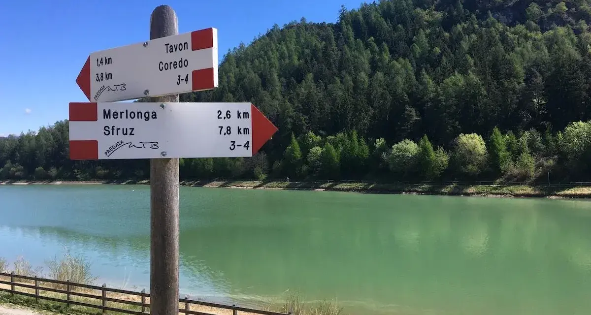 Cartello con indicazioni per Tavon di Coredo, Merlonga e Sfruz, con lago verde e bosco sullo sfondo.