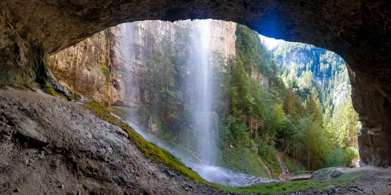 Vista da una grotta su maestose cascate del Trentino, con sentieri e ponticelli che invitano all'esplorazione.