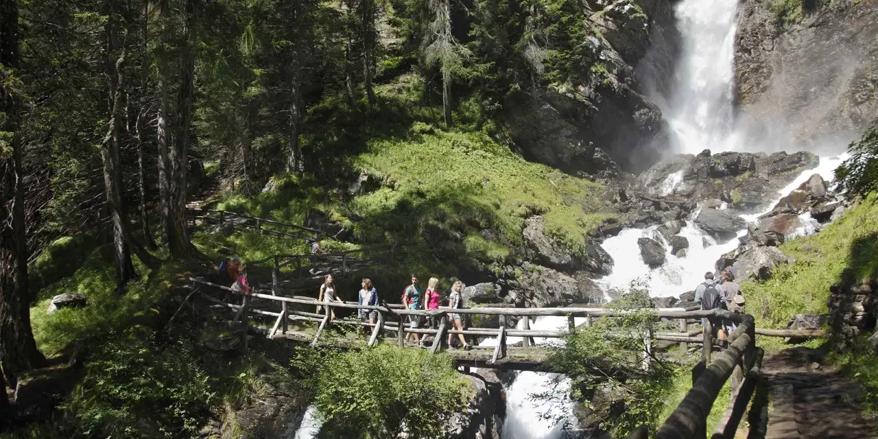 Gruppo di escursionisti attraversa un ponte di legno sul sentiero del Val di Rabbi, con una cascata impetuosa sullo sfondo.