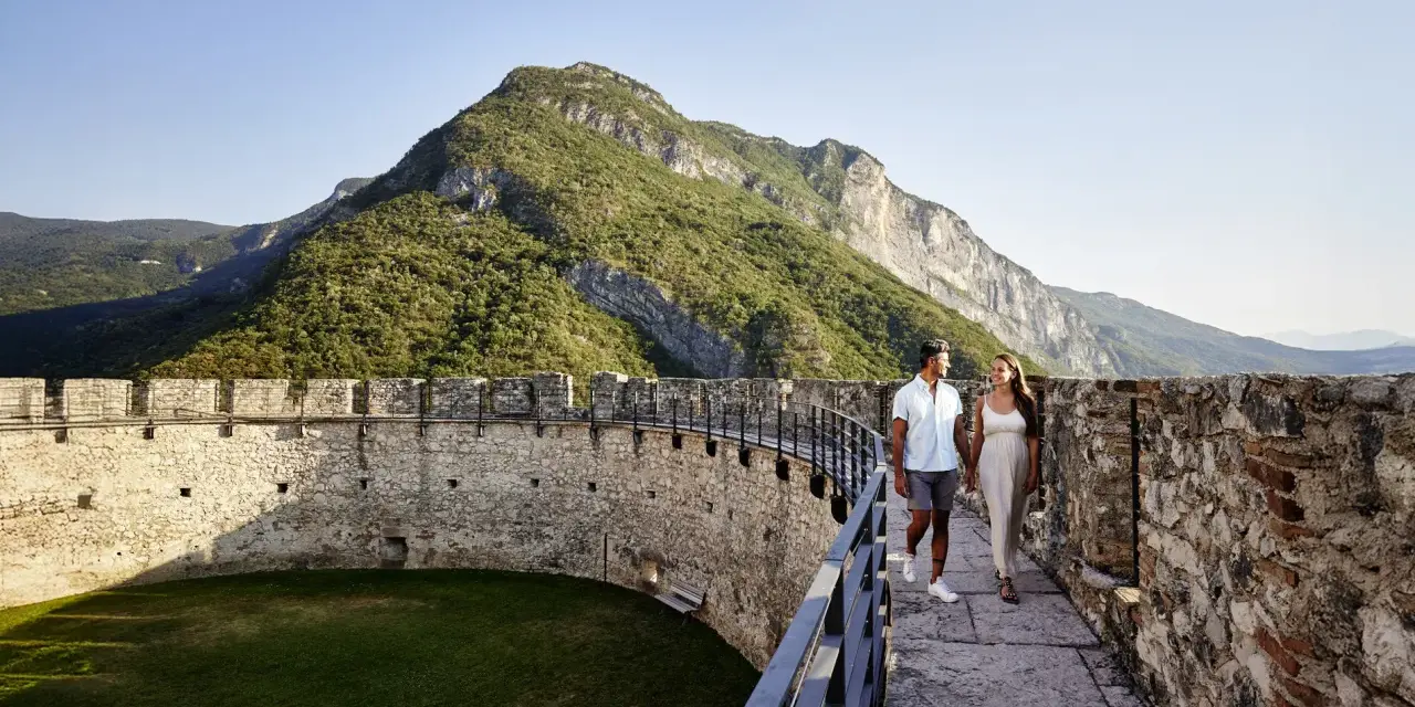 Coppia passeggia su un bastione di un castello con montagne verdi sullo sfondo. Un'esperienza da vivere nel Trentino da scoprire.