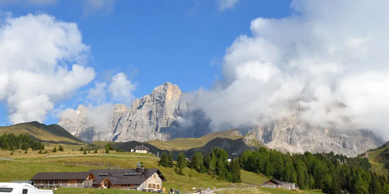 Panorami mozzafiato per escursioni a Passo Rolle. Rifugi alpini e vette imponenti sotto un cielo azzurro con nuvole bianche.