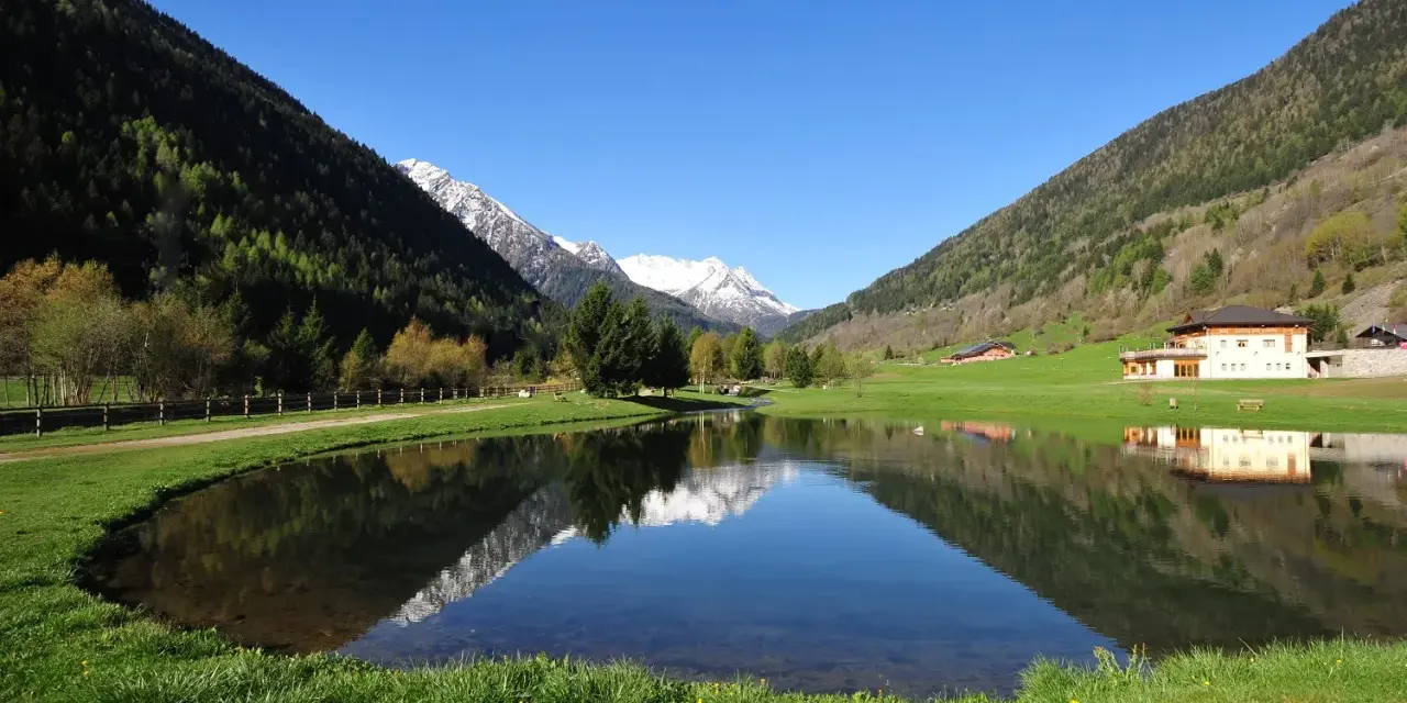 I laghetti di Vermiglio riflettono montagne innevate e un cielo azzurro. Un edificio moderno si specchia nell'acqua calma.