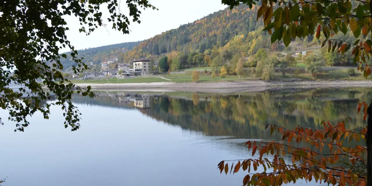 Autunno a Baselga di Pinè: il lago riflette le case e i boschi colorati. Cosa vedere in questo incantevole borgo trentino.