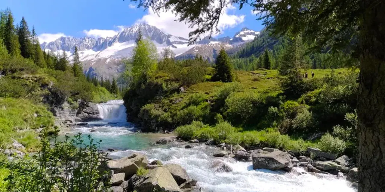 Un torrente impetuoso scorre tra rocce e vegetazione lussureggiante nel **Val di Fumo**, con montagne innevate sullo sfondo.