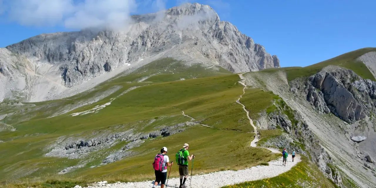 Escursionisti su sentiero ghiaioso, godendosi un **trekking più giorni Dolomiti** con vette imponenti e prati verdi.