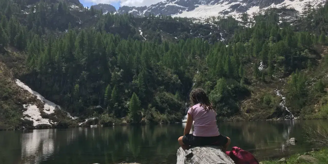 Bambina seduta su un tronco guarda il lago di Barco, circondato da montagne innevate e foreste di conifere.