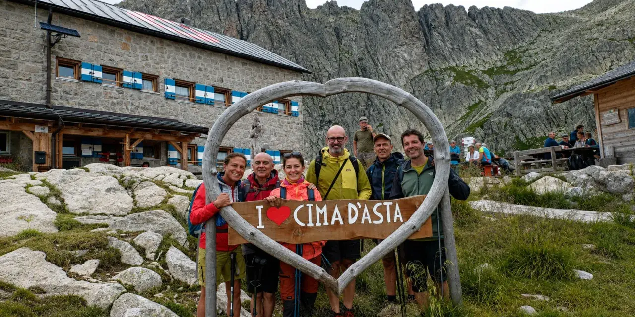 Gruppo di escursionisti felici davanti a un rifugio alpino, con un cartello a forma di cuore che dice "I ❤️ CIMA D'ASTA". Un'indimenticabile esperienza di trekking Trentino.