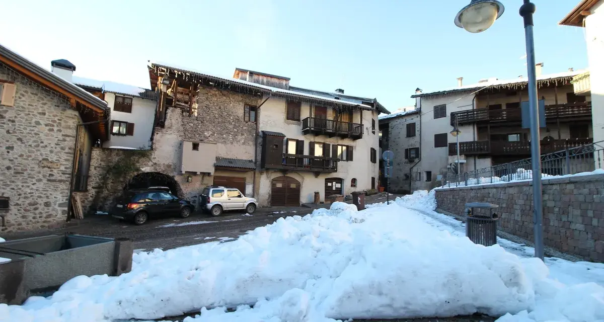 Paesaggio innevato a Miola di Pinè, con case in pietra e legno, auto parcheggiate e un cielo azzurro.