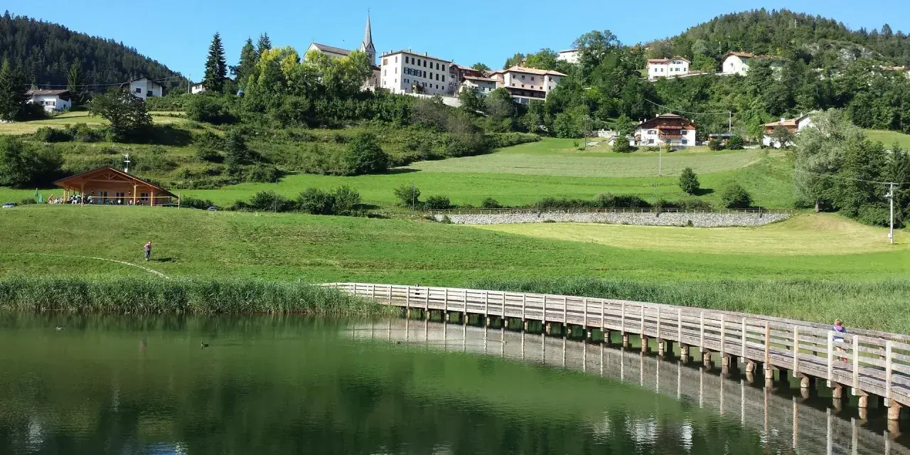Una piacevole passeggiata dria al fos, con un ponte di legno che attraversa il lago e un villaggio sullo sfondo.