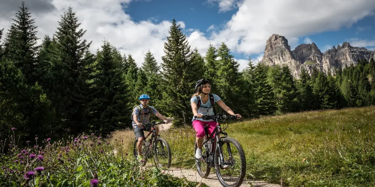 Coppia in mountain bike su sentiero a Passo Campolongo, con bosco di abeti e montagne sullo sfondo.