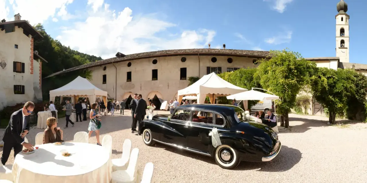 Matrimonio a Caldonazzo centro storico: auto d'epoca nera, tavoli allestiti, ospiti e un campanile sullo sfondo.