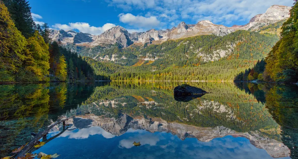 Il **lago di Tovel** riflette montagne maestose e boschi autunnali sotto un cielo azzurro con nuvole bianche. Una roccia emerge dall'acqua cristallina.