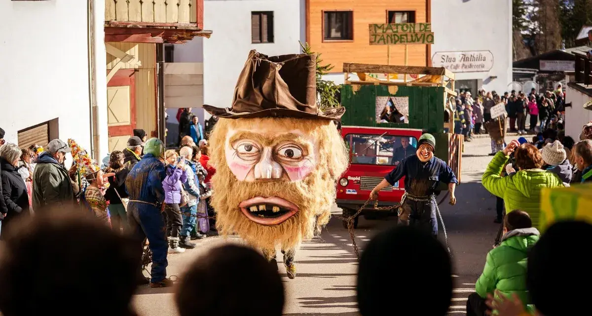Gigantesca maschera pelosa con cappello marrone sfila al carnevale trentino. Un uomo con maschera grottesca trascina una catena.