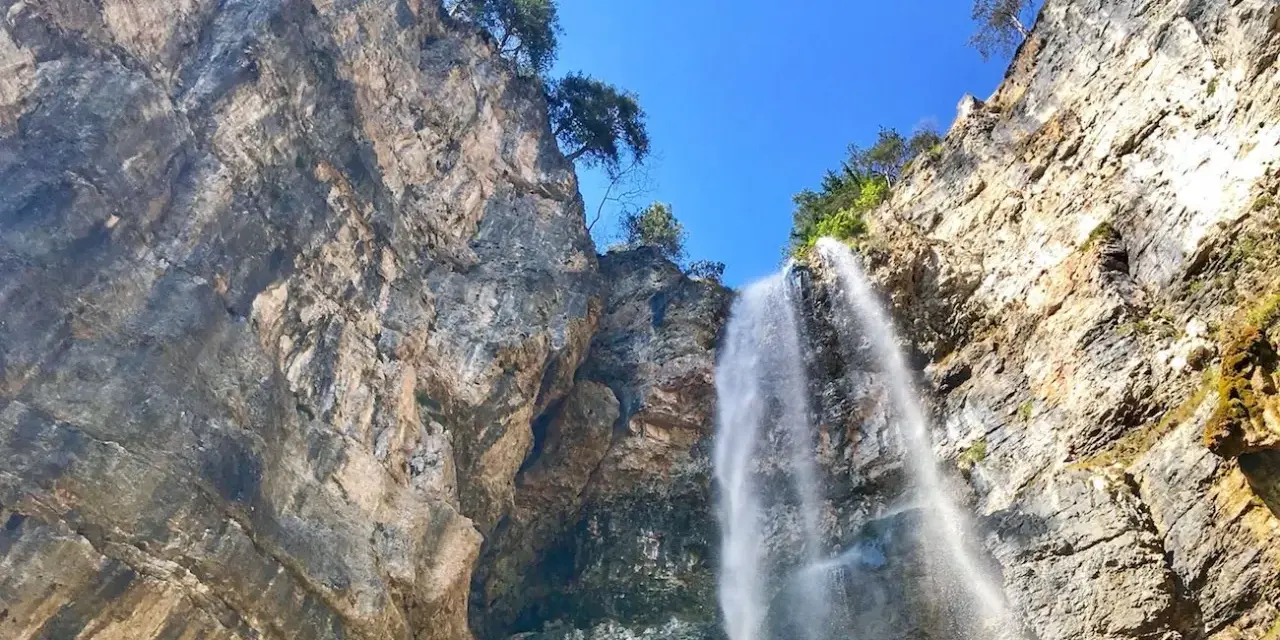 La cascata di Tret scende maestosa da una parete rocciosa imponente, sotto un cielo azzurro e limpido.