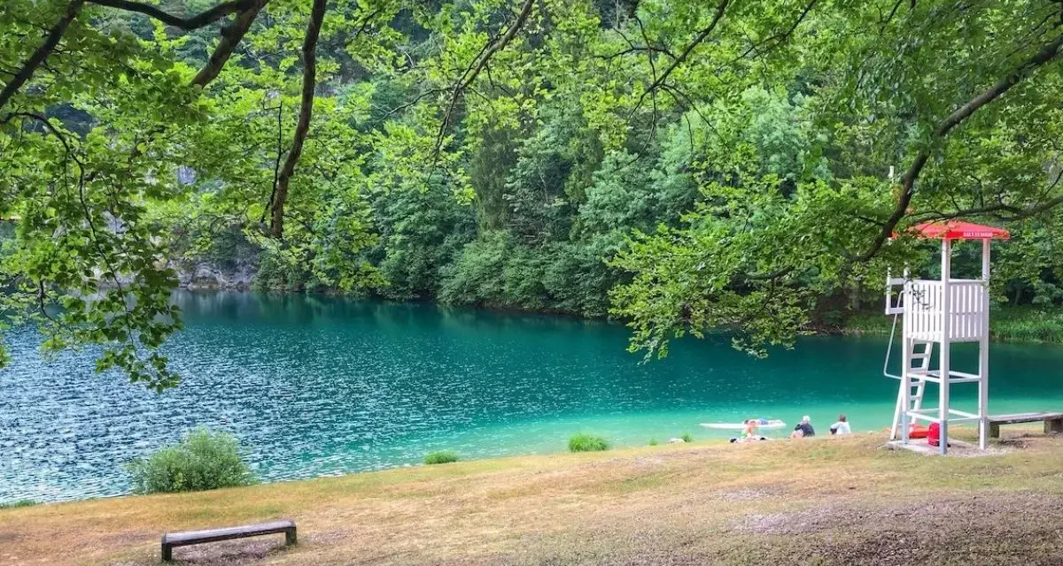 Rilassante **lago Lamar** con acque turchesi, bagnanti e torretta di salvataggio sotto alberi verdi.