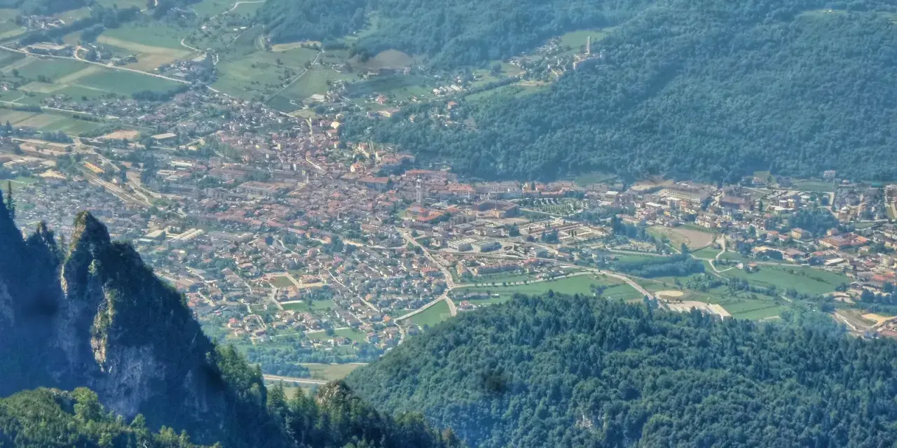 Vista aerea della Valsugana, Trento, con un paese incastonato tra montagne verdi e un picco roccioso in primo piano.