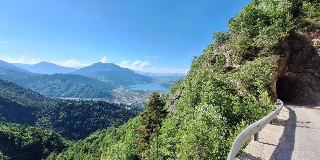 Vista panoramica dal Monte Vezzena con lago azzurro e montagne sullo sfondo. Strada con galleria scavata nella roccia.