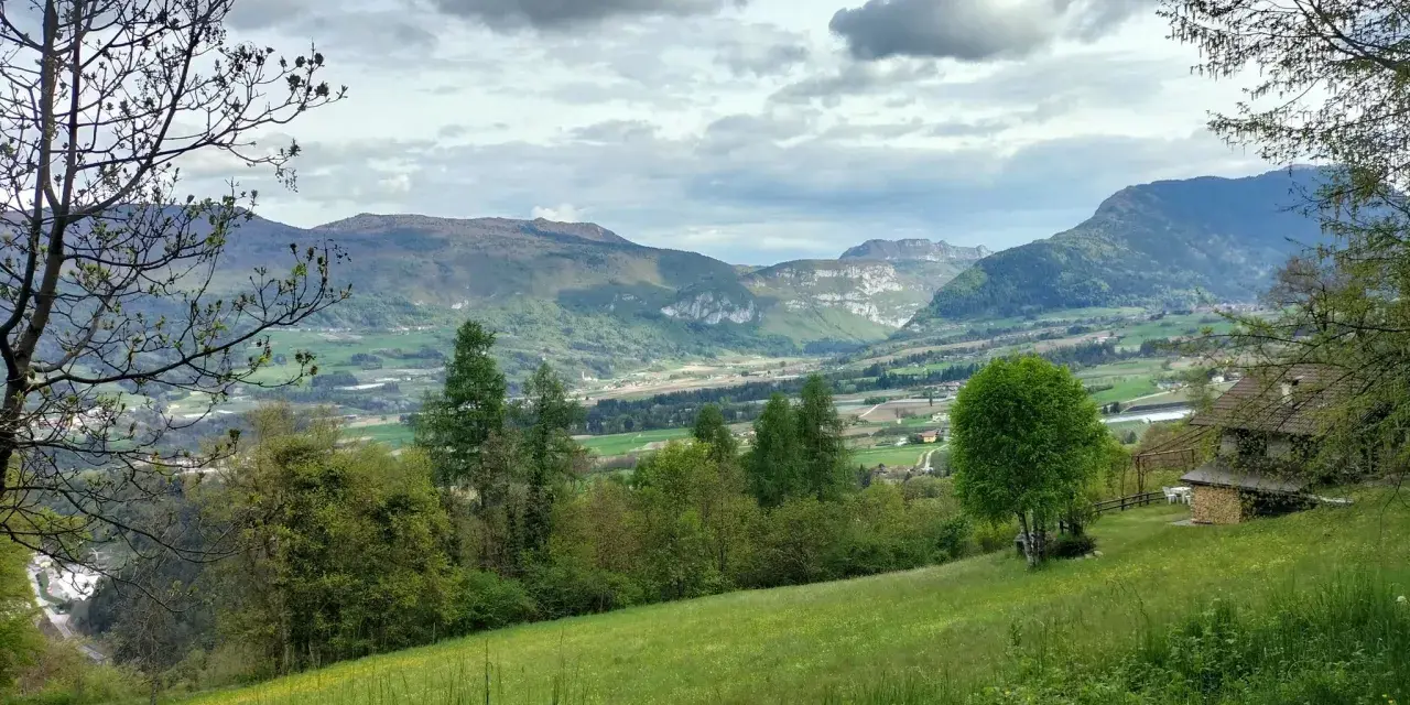 Prato verde e montagne sullo sfondo nella valle di Comano. Una casa con tetto in legno e un albero solitario.