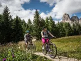 Coppia in mountain bike su sentiero a Passo Campolongo, con bosco di abeti e montagne sullo sfondo.