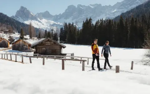 Coppia cammina in una località di montagna trentino inverno, tra baite di legno e montagne innevate.