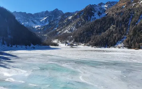 Il lago di Fontana Bianca, un'immensa distesa ghiacciata sotto un cielo azzurro, circondato da montagne innevate e boschi scuri.