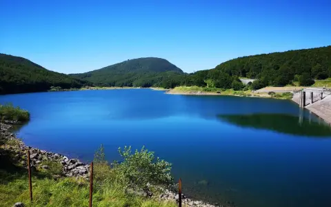 Vasto lago di Comano con acque blu intenso, circondato da colline verdi e una diga in cemento.