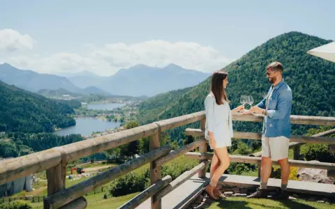 Coppia brinda con vino su un balcone con vista sulla **montagna di Pinè** e laghi.