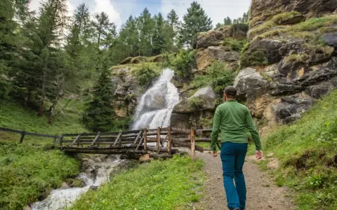 Un escursionista ammira una cascata nel Val di Sole in estate, con un ponte di legno che attraversa il torrente.