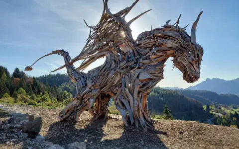 Un maestoso drago di Vaia, scolpito nel legno, domina un paesaggio montano con le ali spiegate verso il sole.