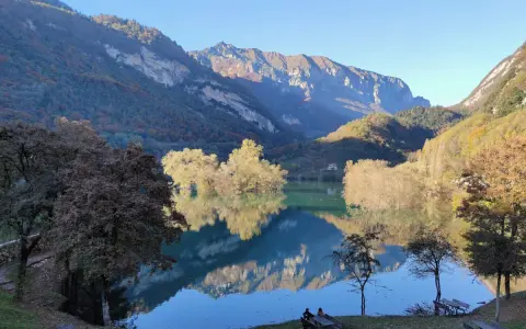 Il lago di Tenno riflette montagne imponenti e alberi autunnali. Due persone siedono su una panchina, godendosi la quiete.
