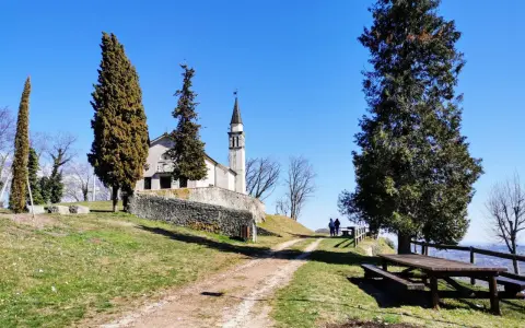A church with a bell tower sits on a grassy hill, with tall trees and a picnic table in the foreground.