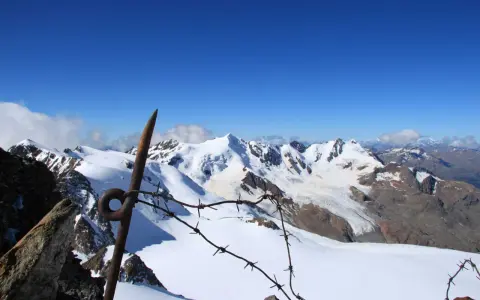 Vista mozzafiato dalla punta Linke, con cime innevate e un cielo azzurro intenso. Un vecchio piccone arrugginito e filo spinato segnano il confine.