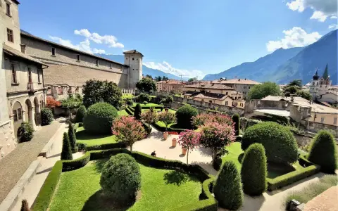 Giardini curati e fioriti incorniciano il maestoso Castel Buonconsiglio, con torri imponenti e montagne sullo sfondo.