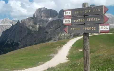 Cartello con indicazioni per Viel del Pan, Passo Fedaia e Passo Pordoi. Sentiero in salita tra prati verdi e montagne imponenti.