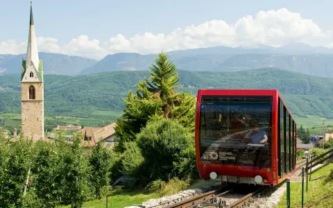 Funicolare rossa in Trentino Alto Adige, con campanile e montagne sullo sfondo. Un'ottima idea per scoprire cosa vedere!
