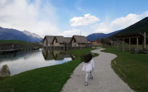 Bambina corre su sentiero ghiaioso verso villaggio palafitticolo nelle Alpi Giudicarie. Montagne sullo sfondo.