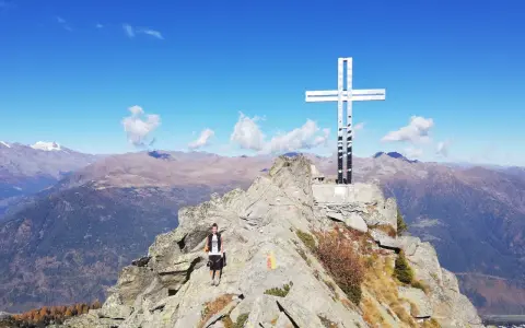Escursionista raggiunge la vetta con la croce della pace, circondata da montagne maestose sotto un cielo azzurro.