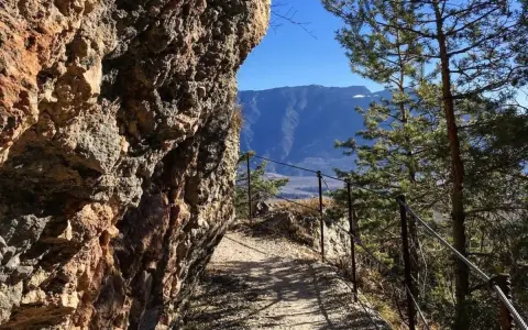 Sentiero margherita: un cammino roccioso con parapetto metallico, alberi e montagne sullo sfondo.