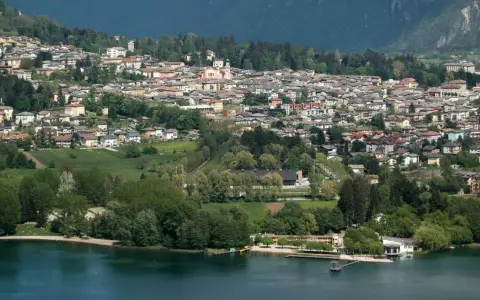 Vista panoramica del **lago di Levico** con il paese arroccato sulla collina. Alberi verdi e case colorate si specchiano nell'acqua tranquilla.