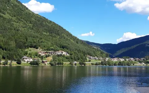 Il **lago di Serraia** riflette le montagne boscose e un villaggio alpino sotto un cielo azzurro con nuvole bianche.
