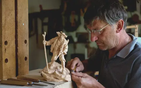 Maestro intaglia un pastore con bastone, un esempio di artigianato legno trentino.