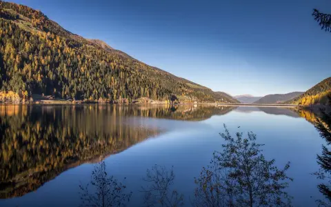 Autunno dorato sul **Val d'Ultimo lago**, con boschi che si specchiano nelle acque calme e un cielo azzurro intenso.